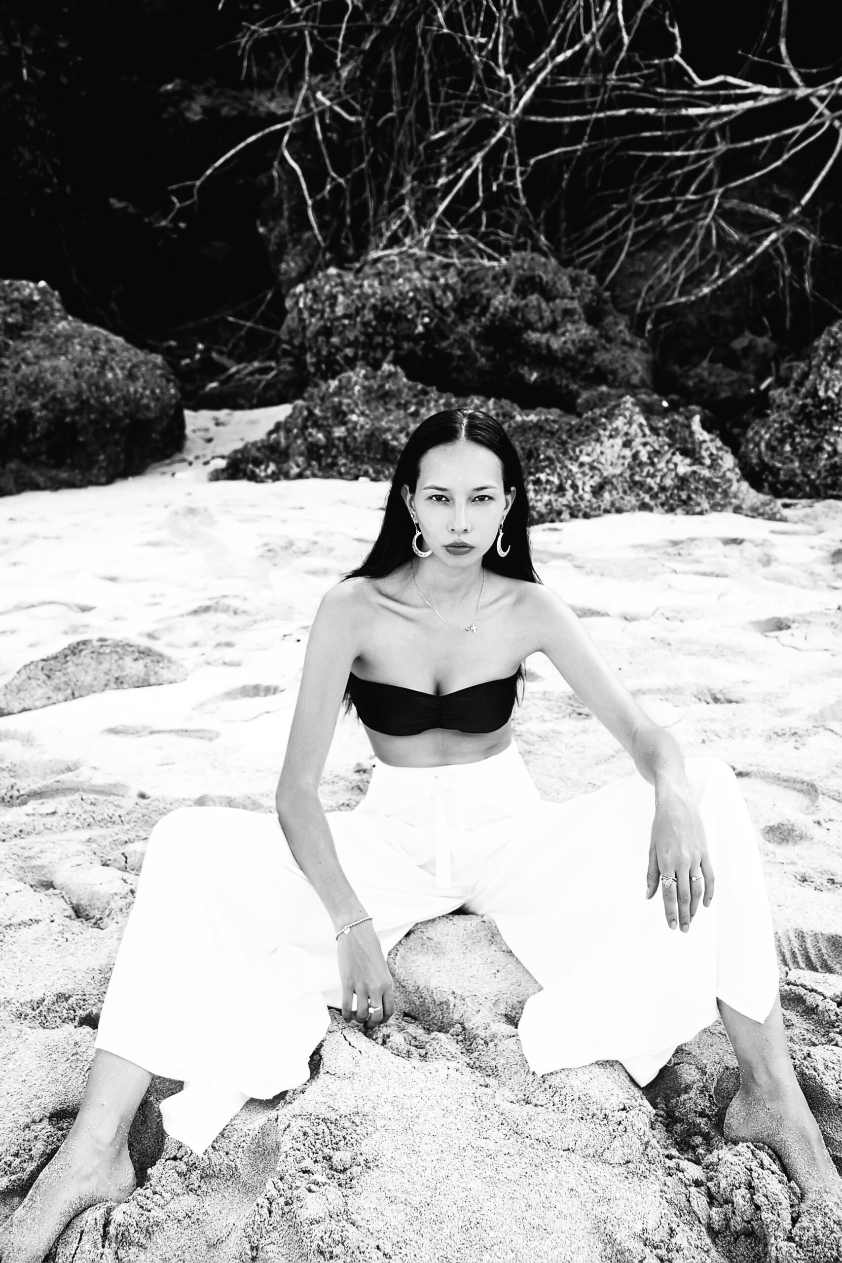 portrait of beautiful caucasian woman model with dark long hair in wide-leg classic pants  sittting on summer beach with white sand near rocks